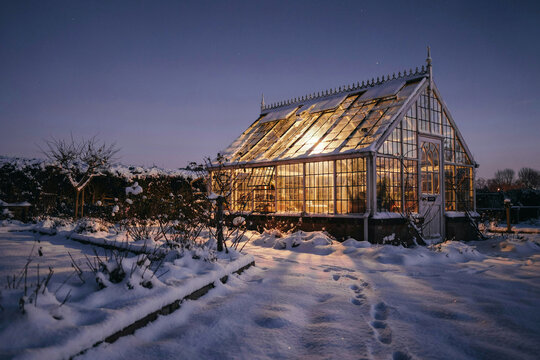 Glass greenhouse glowing at dusk in snowy winter garden, warm interior lights and frost-covered paths, tranquil rural scene for seasonal nature concept