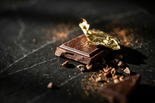 Dark chocolate bar with gold foil and cocoa beans on black slate background, rustic food styling and moody lighting for dessert or gourmet packaging