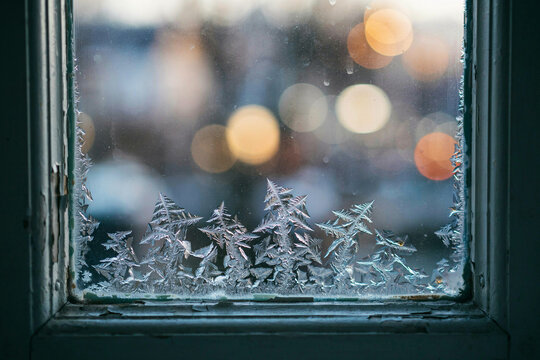 Frosted window glass with ice crystals and warm city bokeh lights, winter cold weather background for seasonal design and holiday promotions