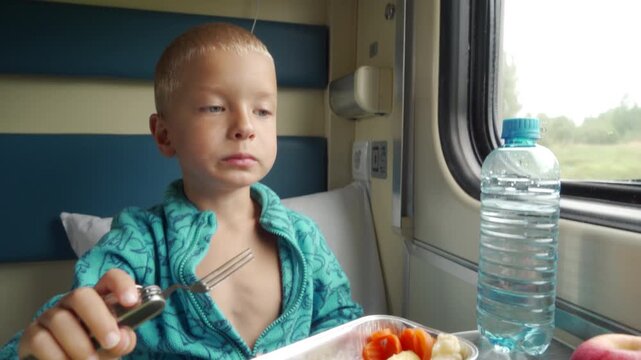 A happy boy eats his lunch while riding inside a cozy train compartment. His playful and curious look brings a warm, joyful feeling to the quiet travel day.