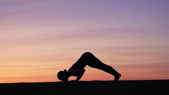 Silhouette of woman holding inverted yoga pose on open ground with pastel sunset sky, low horizon, calm backlit profile and strong graceful balance in outdoor wellness scene