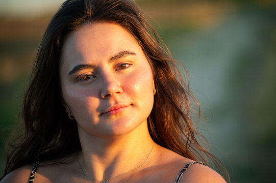 Close up portrait of a young woman with long brown hair looking at the camera. The scene is illuminated by warm golden hour sunlight with a blurred natural background.
