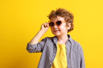 Fototapeta premium Cute little boy in stylish sunglasses and checkered shirt posing on yellow background