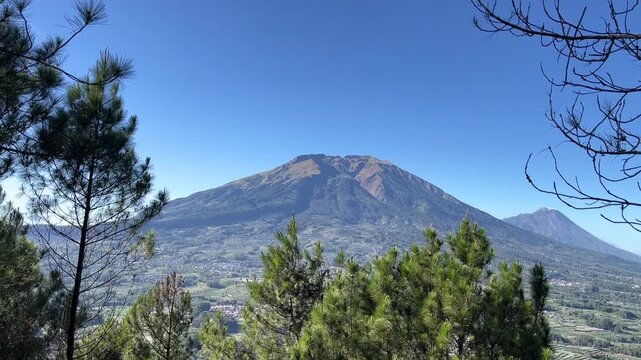 Scenic landscape view of Mount Merbabu under a clear blue sky, showing green slopes, terraced agricultural fields, and rural village settlements.