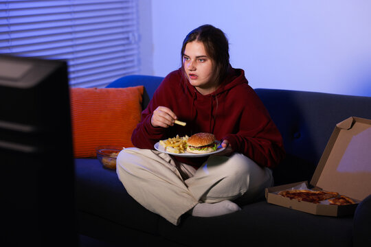 Overweight woman eating French fry and watching TV on sofa at home