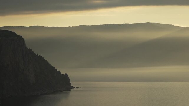 Mountain coastline mist at sunrise with sunbeams filtering through clouds over a calm sea in a scenic landscape