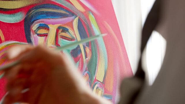 Close-up of an artist&rsquo;s hand painting a colorful abstract portrait on canvas with a brush. Creative process in a bright art studio showcasing vibrant acrylic or oil painting techniques.
