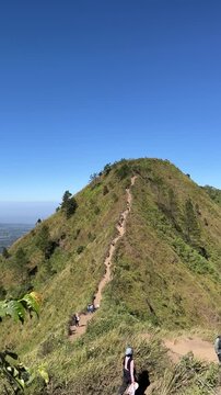 Panoramic view of Mount Andong peak with hikers along the grassy ridge trail under a clear blue sky, showing surrounding valleys and lush green slopes.