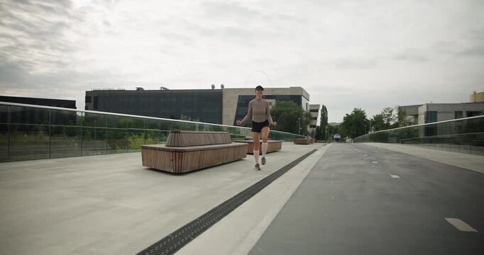 A young beautiful girl in sportswear and a cap does exercises with a skipping rope and plays sports in an urban setting on a bridge with a pedestrian area and benches against the backdrop of new concr