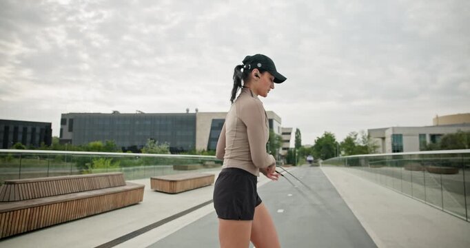A young beautiful girl in sportswear and a cap does exercises with a skipping rope and plays sports in an urban setting on a bridge with a pedestrian area and benches against the backdrop of new concr