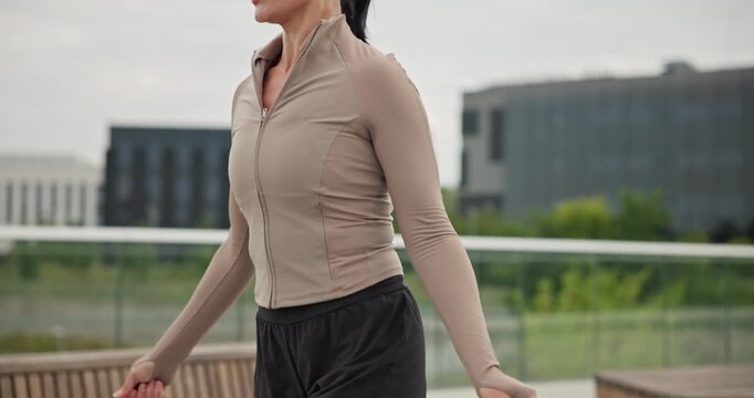 A young beautiful girl in sportswear and a cap does exercises with a skipping rope and plays sports in an urban setting on a bridge with a pedestrian area and benches against the backdrop of new concr