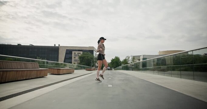 A young beautiful girl in sportswear and a cap does exercises with a skipping rope and plays sports in an urban setting on a bridge with a pedestrian area and benches against the backdrop of new concr