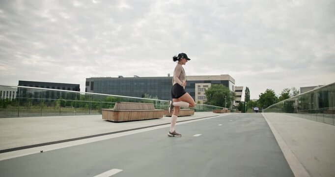 A young beautiful girl in sportswear and a cap does exercises with a skipping rope and plays sports in an urban setting on a bridge with a pedestrian area and benches against the backdrop of new concr