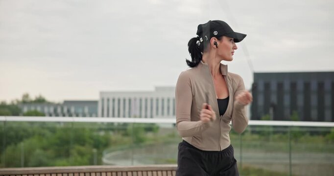 A young beautiful girl in sportswear and a cap does exercises with a skipping rope and plays sports in an urban setting on a bridge with a pedestrian area and benches against the backdrop of new concr