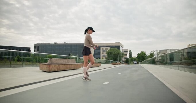 A young beautiful girl in sportswear and a cap does exercises with a skipping rope and plays sports in an urban setting on a bridge with a pedestrian area and benches against the backdrop of new concr