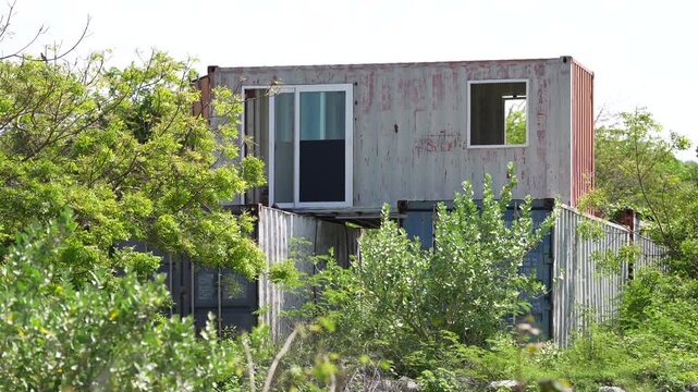 Shipping container modular home container house rural overgrown abandoned window sliding door weathered metal structure, recycled off grid eco housing amid green vegetation