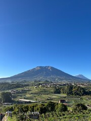 Obraz premium Scenic landscape view of Mount Merbabu under a clear blue sky, showing green slopes, terraced agricultural fields, and rural village settlements.