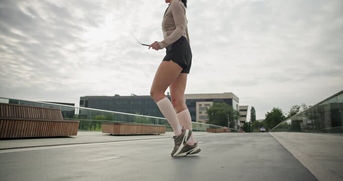 A young beautiful girl in sportswear and a cap does exercises with a skipping rope and plays sports in an urban setting on a bridge with a pedestrian area and benches against the backdrop of new concr