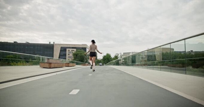A young beautiful girl in sportswear and a cap does exercises with a skipping rope and plays sports in an urban setting on a bridge with a pedestrian area and benches against the backdrop of new concr