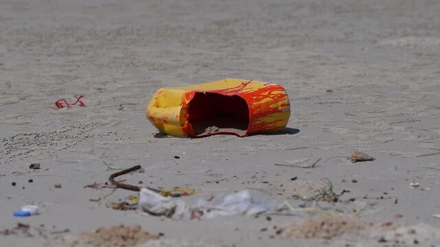 Beach sand plastic litter pollution waste environment debris closeup of crushed container and discarded packaging on shoreline, broken yellow canister amid scattered waste, urgent cleanup warning