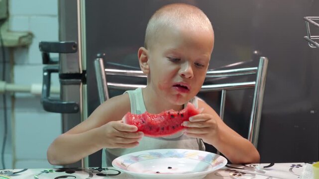 A happy little boy eagerly eats a juicy slice of watermelon at the kitchen table. He looks down with satisfaction inside his cozy home.