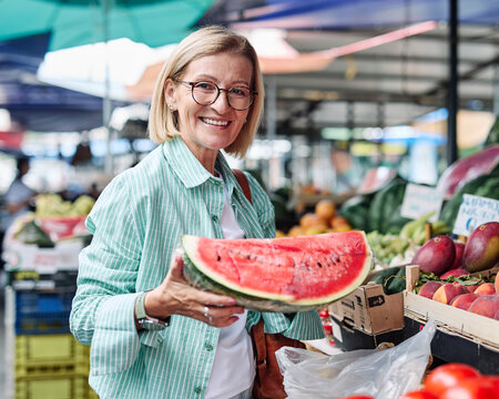 A woman enjoys selecting ripe tomatoes at a bustling outdoor farmers market. Colorful fruits and vegetables surround her as she smiles at the freshness of the produce