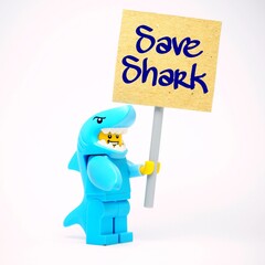Fototapeta premium Indonesia - October 4, 2025 : Mini toy of action figure at table with blurred background. Miniature photography concept design. Minifigure of Shark guy character hold SAVE ME poster banner campaign