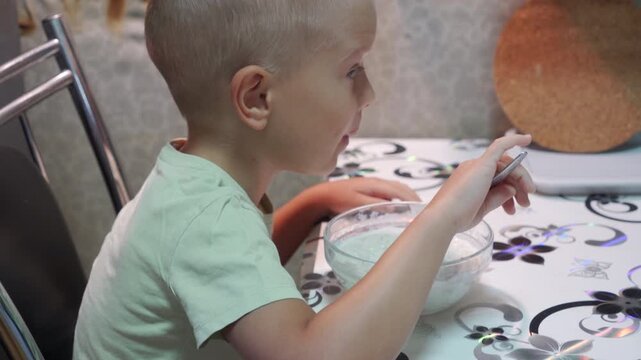 A calm little boy happily eats his morning porridge with a spoon. He sits contentedly at a patterned kitchen table inside his home.