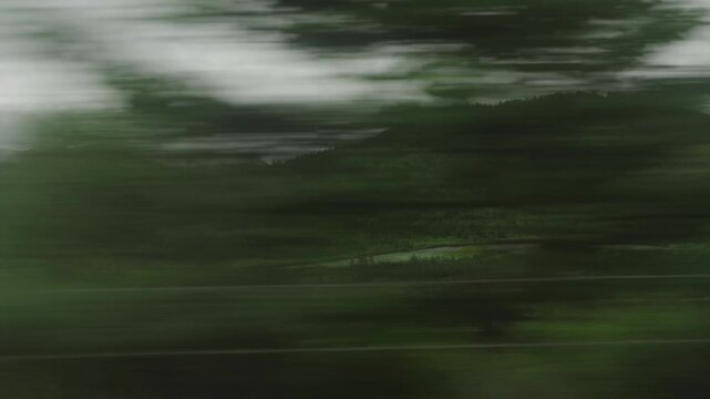 A train swiftly moves through lush green hills under an overcast sky, capturing rolling landscapes dotted with trees and small structures along its path.