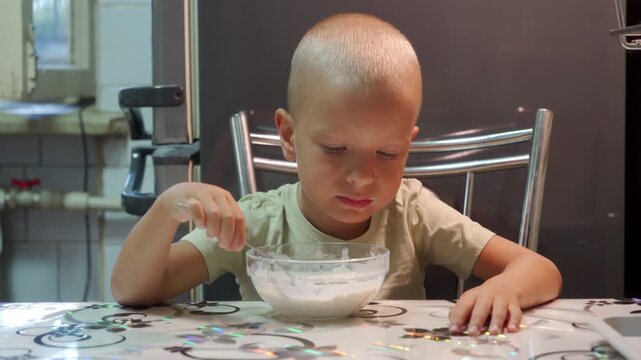 A serious little boy holds a spoon over a bowl of porridge. He looks directly forward at a kitchen table, feeling focused and intense during his breakfast.
