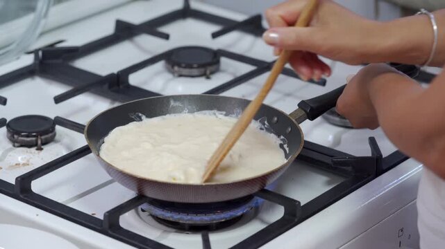 A focused cook gently stirs a creamy white sauce in a frying pan. She prepares a comforting meal on a gas stove inside a cozy kitchen.