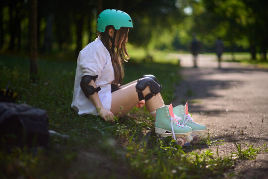 Girl sits on the ground in a park while wearing roller skates and safety gear in the afternoon light