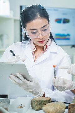Scientist laboratory sample tablet safety goggle glove stone specimen analysis, focused Asian female researcher studies mineral material during clean experiment
