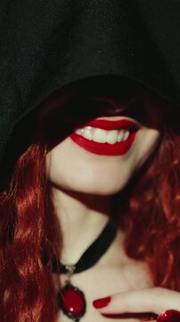 Close-up mouth red lips beauty face sexy vampire woman happy smiling shows white long false fangs. art halloween party make-up. Gothic lady girl hiding face black hood dress vertical video crop short.