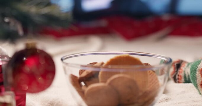 A close-up of a glass bowl filled with cookies next to feet in warm patterned knitted socks on a soft throw.