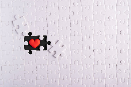 Top view of white jigsaw puzzle with one missing piece revealing a red heart on black background. Concept of finding true love, soulmate, passion, completion, and romance.