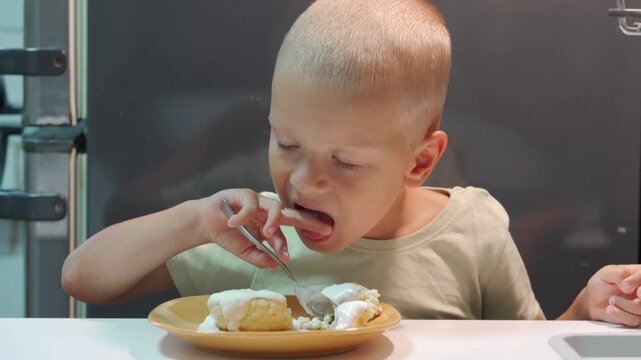 A hungry little boy happily licks sweet white cream from his finger while eating pancakes with a spoon. He sits comfortably at a bright table inside a cozy, modern kitchen.