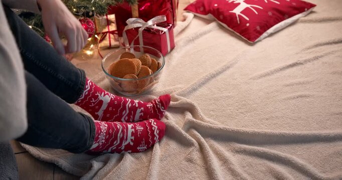 A person reaches for a bowl of cookies while relaxing in red patterned Christmas socks near a tree with wrapped gifts.