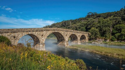 Fototapeta premium Stone bridge over a river with lush greenery and blue sky.