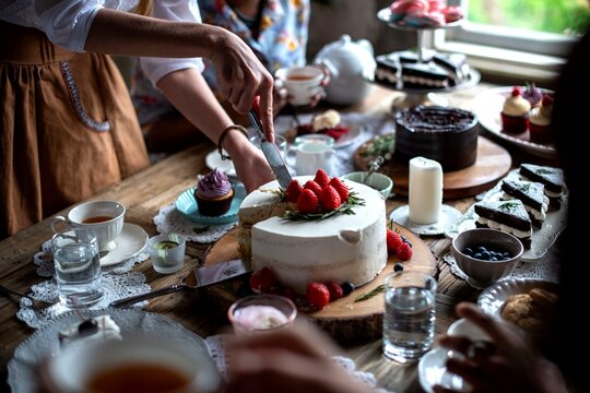 Hands cutting a cake on a rustic table with various desserts and tea.