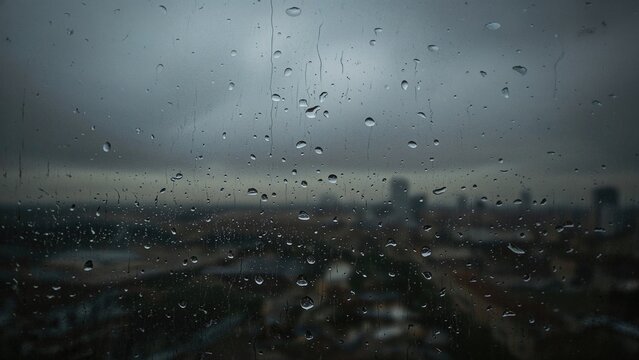 Rainy window with water droplets, blurry cityscape outside, overcast sky, gloomy weather, urban view through glass.