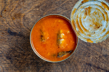 Naklejka na ściany i meble Top down view of an open metal tin can filled with fish in bright orange tomato sauce placed on rustic wooden table, closeup