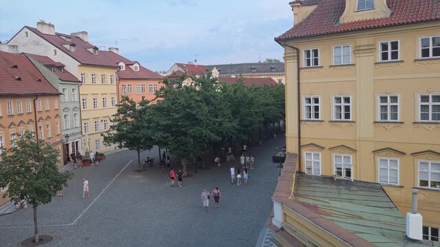 Panning shot of pedestrians strolling through historic cobblestone square with trees and pastel buildings.  Tourists in central Prague.