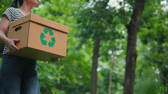 Young Asian woman volunteer holding a recycling box in the park. Concept of recycling, environmental protection, sustainability, zero waste, and ESG strategy on green natural forest background.