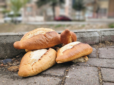 Pile of fresh bread loaves discarded on a paved city sidewalk next to a curb, with a blurred urban street background