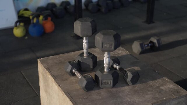 Woman wearing a watch reaching for and picking up a water bottle placed beside hex dumbbells on a wooden plyometric box in a modern indoor gym.