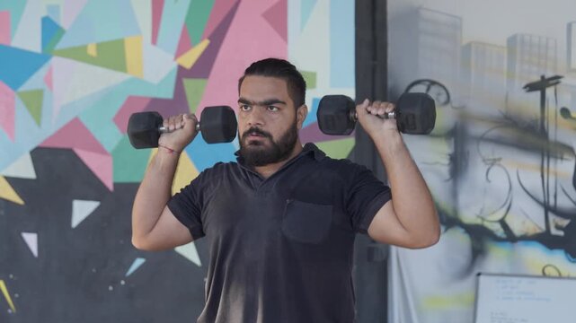 Bearded South Asian male athlete performing a standing dumbbell shoulder press during a strength training workout in a modern indoor fitness studio. Camera moving in a mid close arc around the subject
