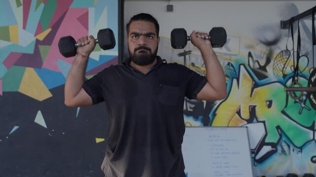 Bearded South Asian male athlete performing a standing dumbbell shoulder press during a strength training workout in a modern indoor fitness studio. Camera tracks in and then tracks out during the exe