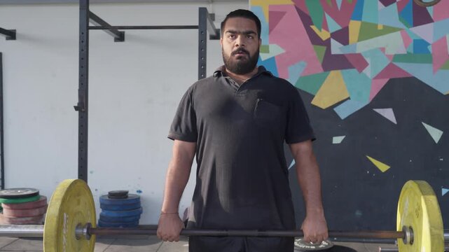 Bearded South Asian male athlete performs a barbell deadlift during functional fitness training while the camera moves smoothly side to side from a front view. Collection: WORKOUTSERIES2026