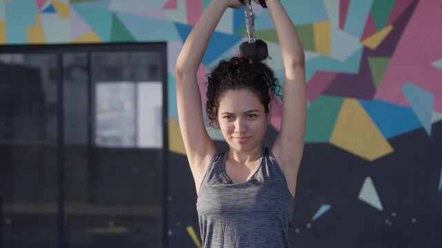 South Asian female athlete performs standing dumbbell overhead tricep extensions. Arc tracking shot with mid close framing. Collection: WORKOUTSERIES2026.